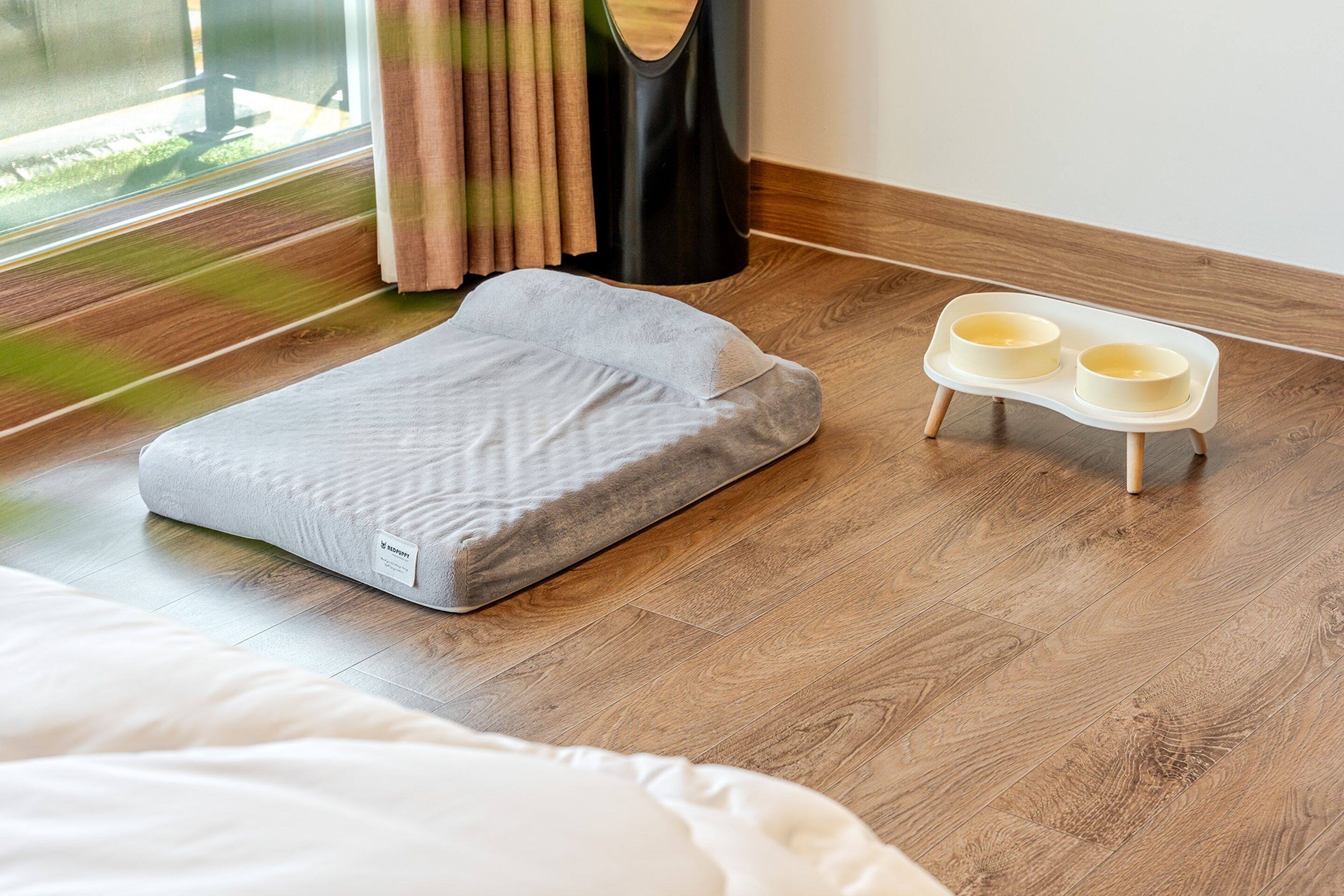 Ein Zimmer mit grauem Bett, einer Tierfutterstelle und einem Fenster mit Vorhängen.
