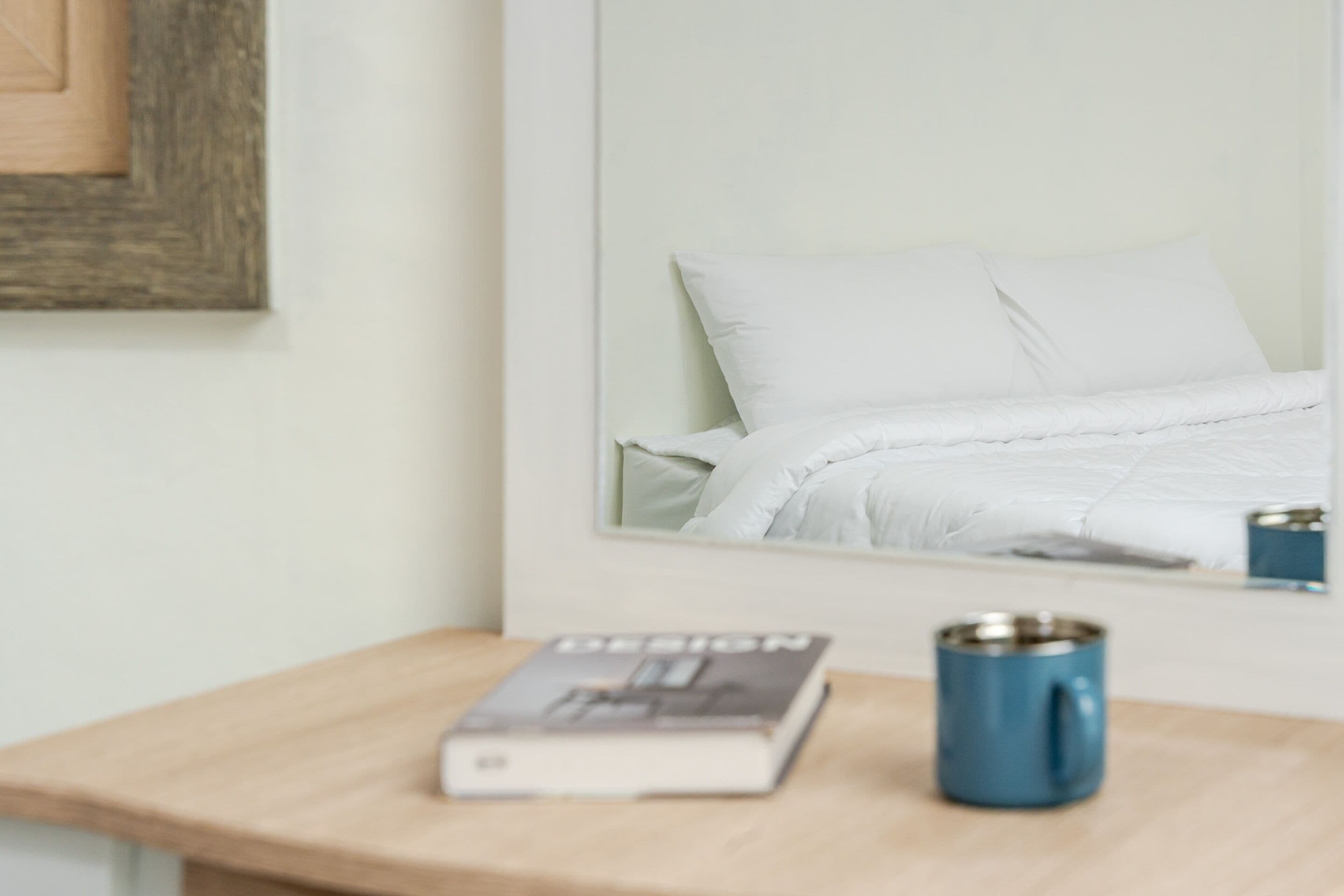 Una cama bien hecha con sábanas blancas, un libro titulado 'DESIGN' sobre una mesita de noche de madera y una taza azul.