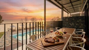 Ein Holztisch mit einem Steak, Gemüse und Weingläsern auf einem Balkon mit Blick auf einen Pool und einen Sonnenuntergang.