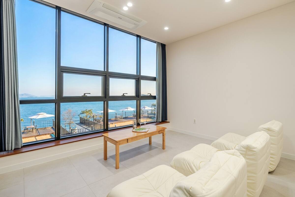 A modern room with large windows, a wooden table, and white upholstered furniture.