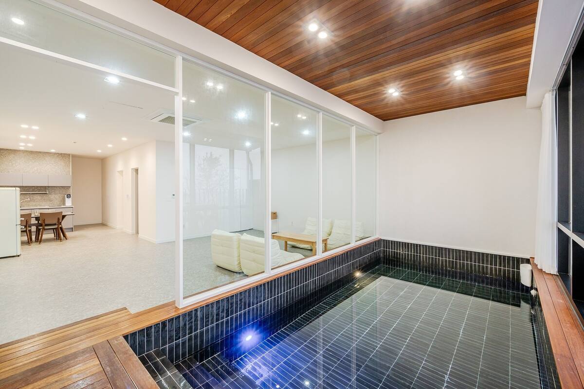 A modern indoor swimming pool with a wooden ceiling, a glass wall, and a seating area with cushions.