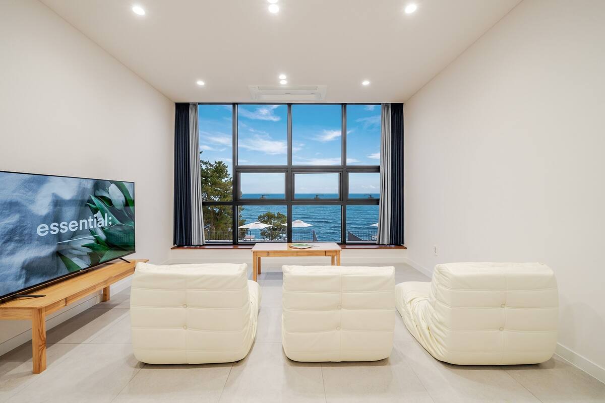 A modern living room with a large TV, three white cushioned chairs, a wooden coffee table, and a view of the sea through large windows.
