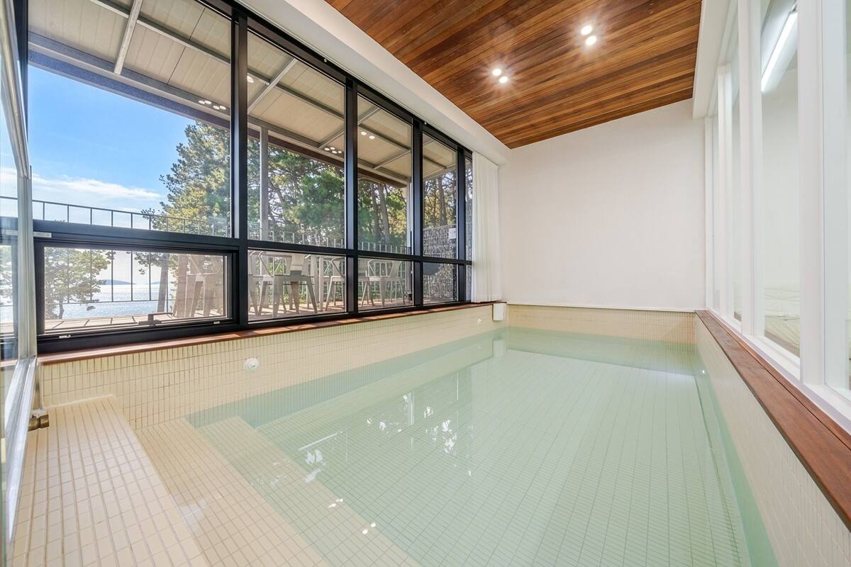 An indoor swimming pool with clear glass walls and a tiled floor, large windows, and a wooden ceiling.