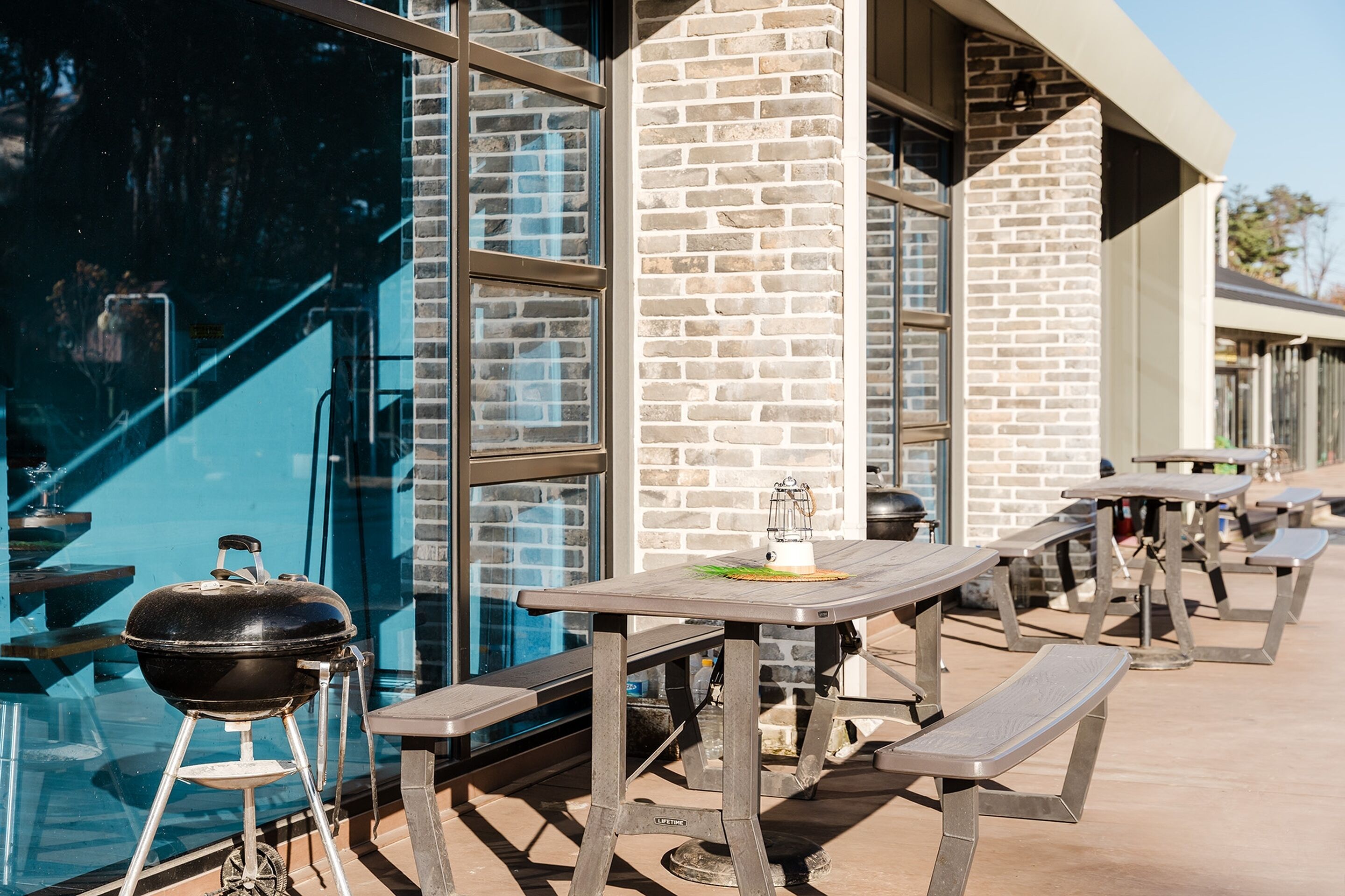Mesas de comedor al aire libre y una parrilla de barbacoa frente a un edificio con ventanas grandes y paredes de ladrillo.