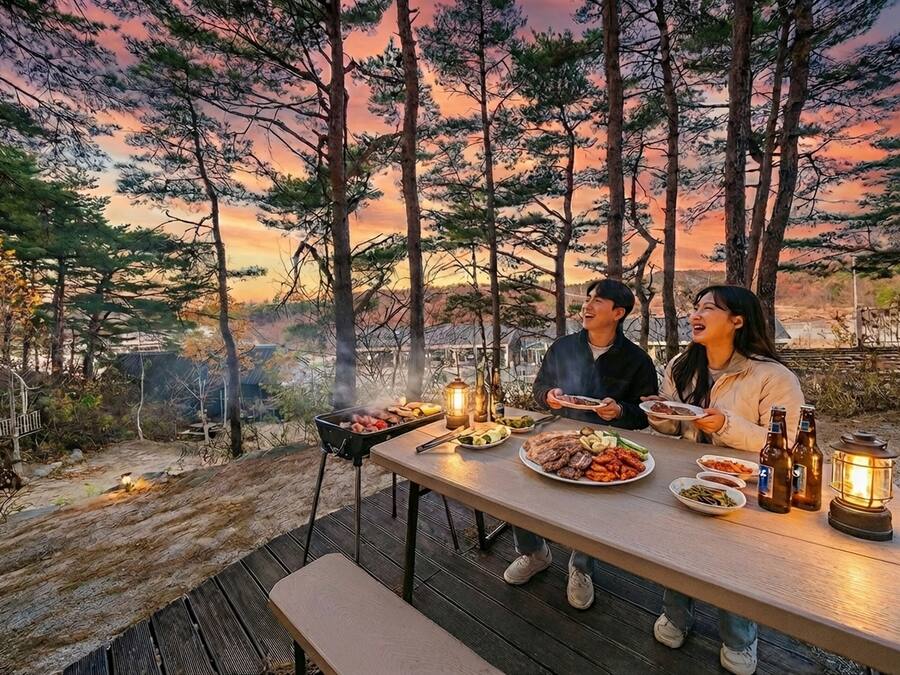 A dining table with food and drinks, set against a forest backdrop with a sunset.