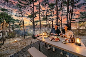 A dining table with food and drinks, set against a forest backdrop with a sunset.