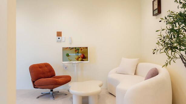 Een moderne kamer met een bruine stoel, een witte bank en een klein tafeltje. Aan de wand hangt een televisie die een afbeelding toont met het woord 'essential' en fruit.