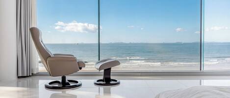 Een moderne kamer met een groot raam met uitzicht op een strand, een ligstoel en een kleine tafel.