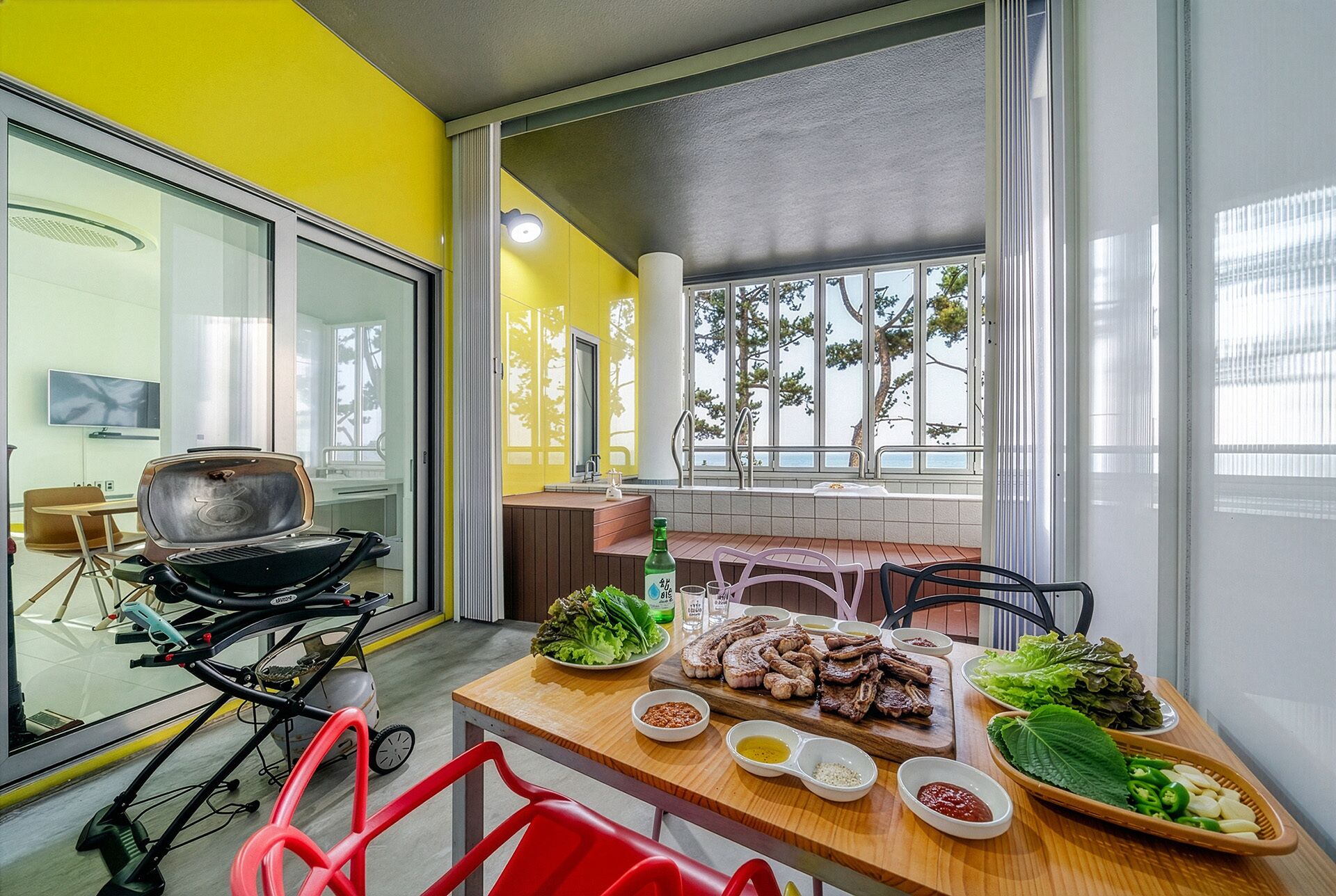 Ein Essbereich mit Grill, einem gedeckten Tisch mit Speisen und Blick auf Bäume durch große Fenster.