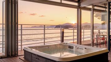 A hot tub on a wooden deck with a view of the ocean and mountains at sunset.