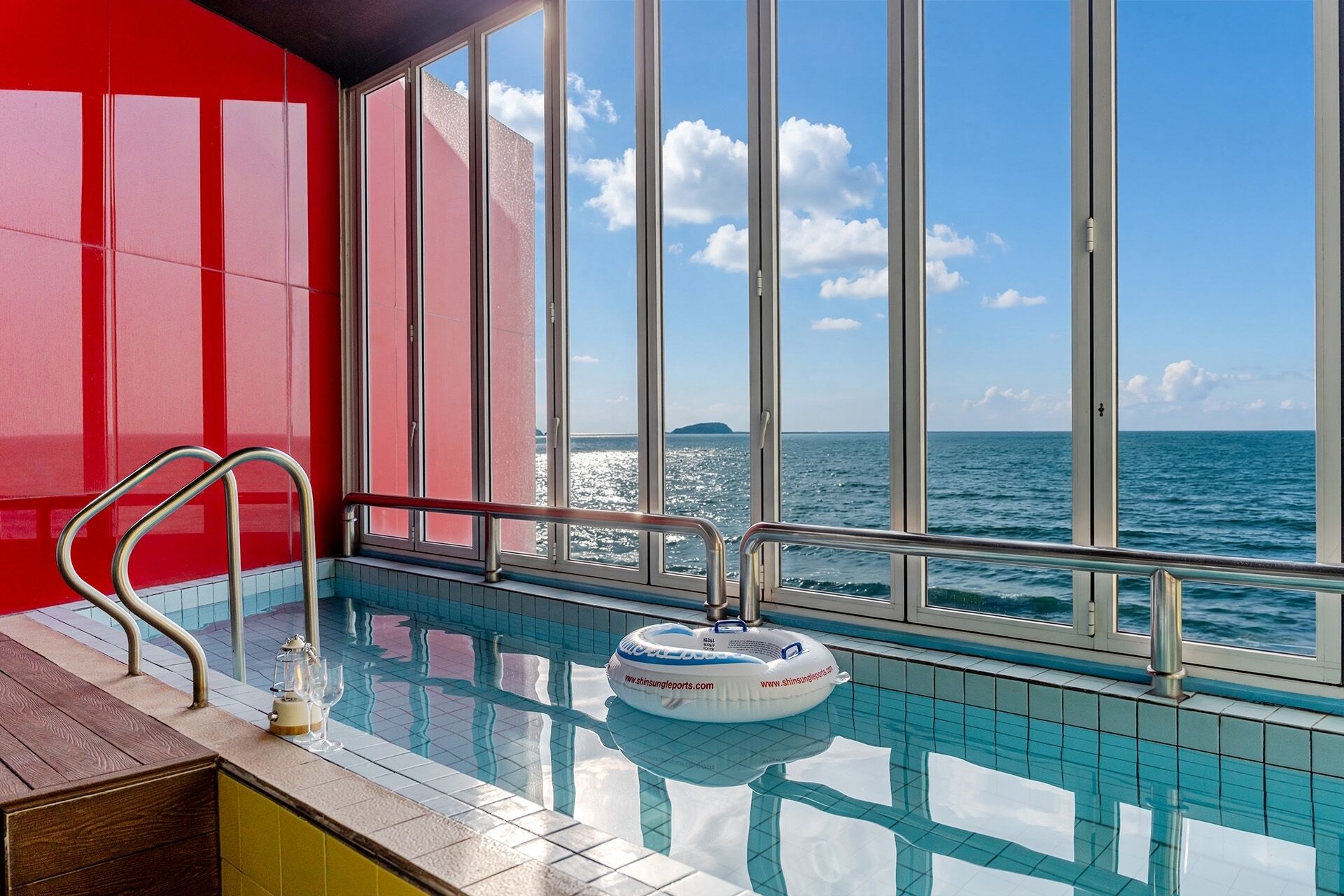 A pool with a clear view of the sea and a distant island, a red wall, and a wooden bench.