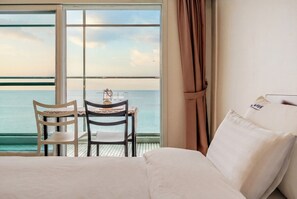 Ein Hotelzimmer mit großem Fenster und Meerblick, einem Bett mit weißer Bettwäsche, einem Tisch mit zwei Stühlen und einem Vorhang.