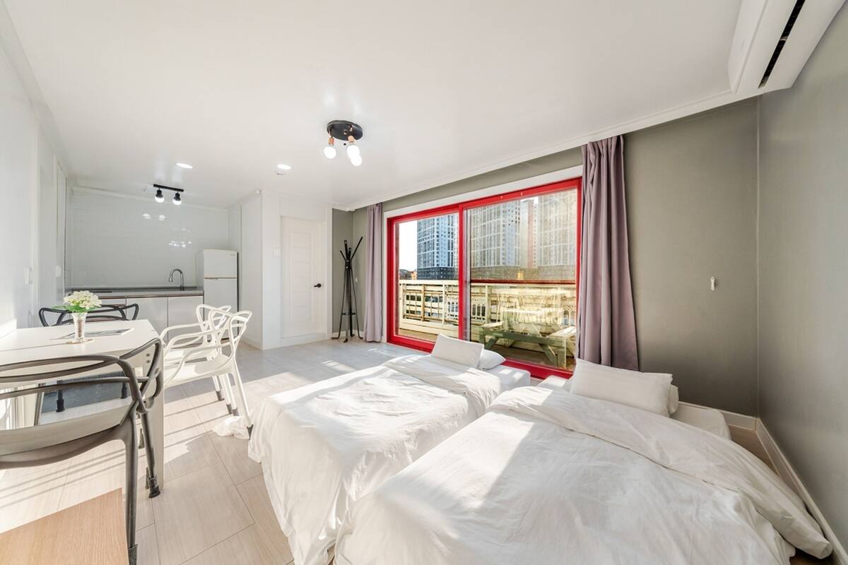 A modern hotel room with a large bed, a dining area with a table and chairs, and a view of a cityscape through a large window.
