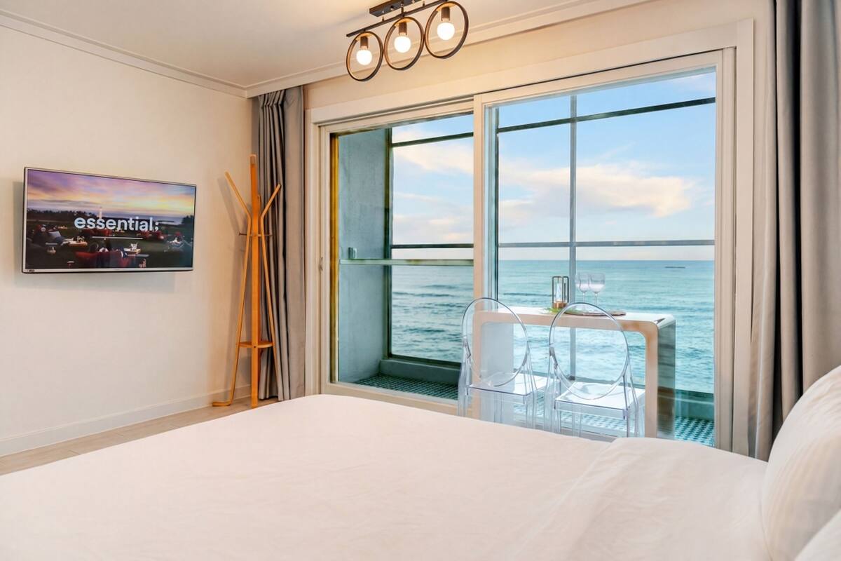 A hotel room with a large bed, a TV on the wall, a window with a view of the sea, and a table with chairs outside the window.