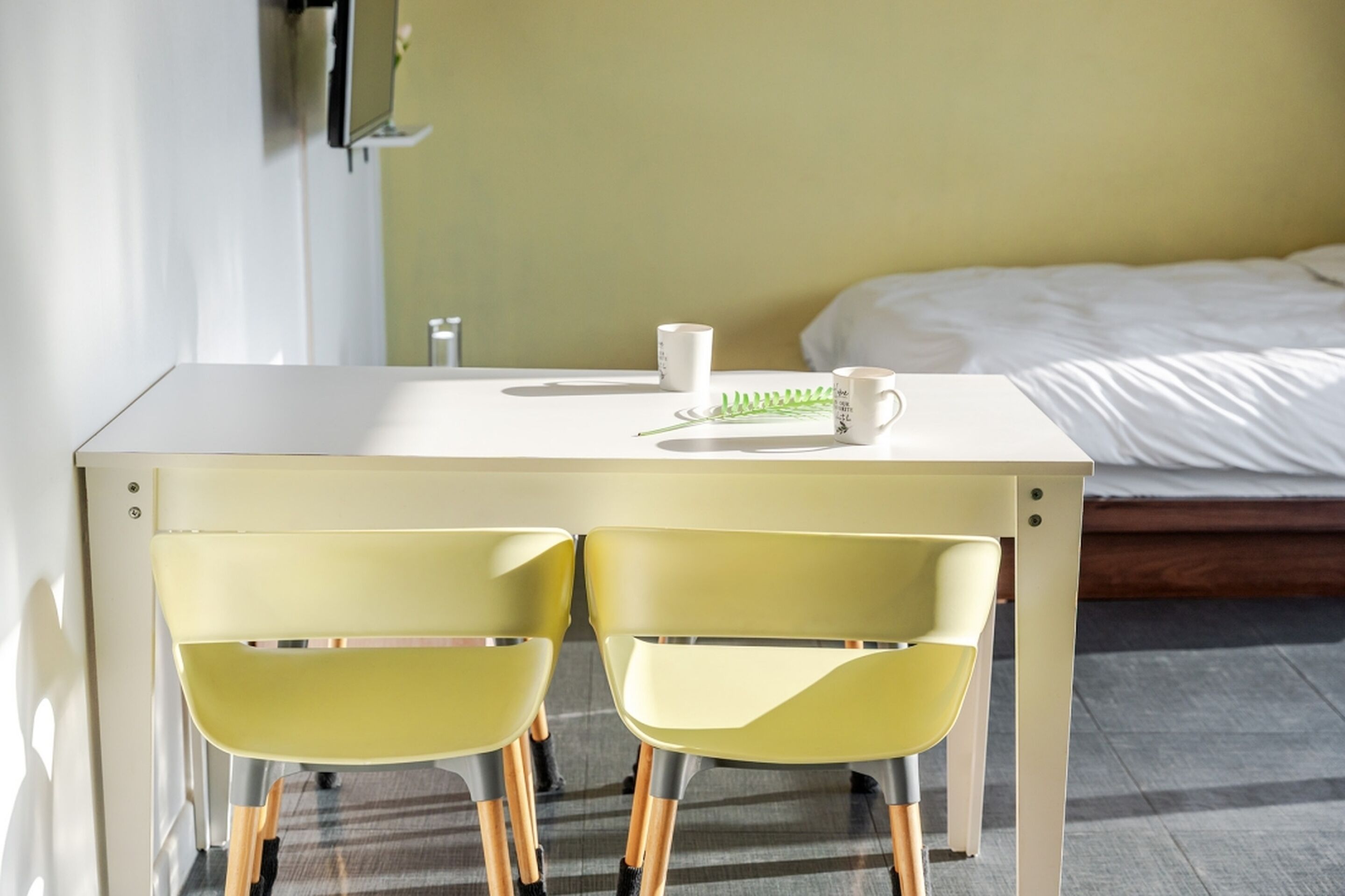 A small, modern room with a single bed, a desk with two chairs, and a bedside table with a cup and a plant.