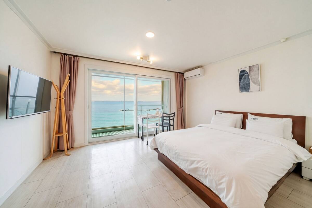 A hotel room with a large bed, a TV, a desk with a chair, and a view of the sea.
