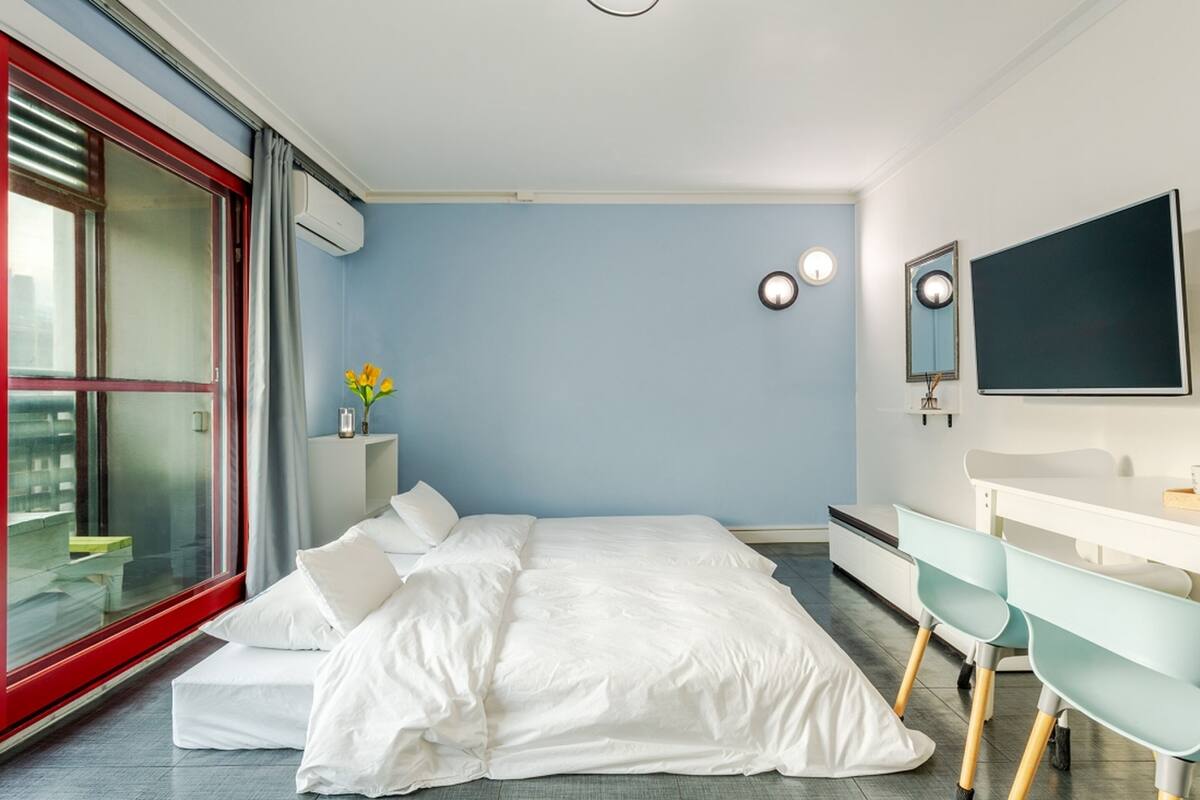 A modern bedroom with a large bed, a sliding glass door, a white bedside table, a mirror, and a flat-screen TV.