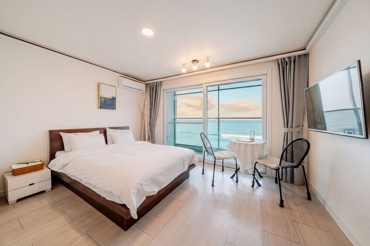 A modern hotel room with a large bed, a small table, two chairs, a TV, and a view of the sea through a large window.