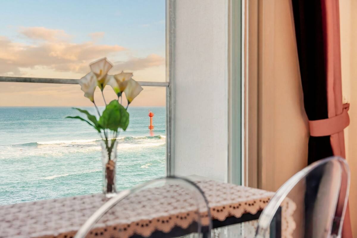 A table with a lace tablecloth and a vase with white flowers, overlooking the ocean with a view of the sea and a buoy.