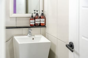 Un baño con un lavabo blanco, un espejo encima y tres frascos de artículos de tocador en un estante.