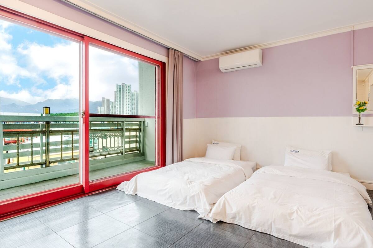 A hotel room with two beds, a large window with a view of a cityscape and a bridge, and a sliding glass door leading to an outdoor balcony.