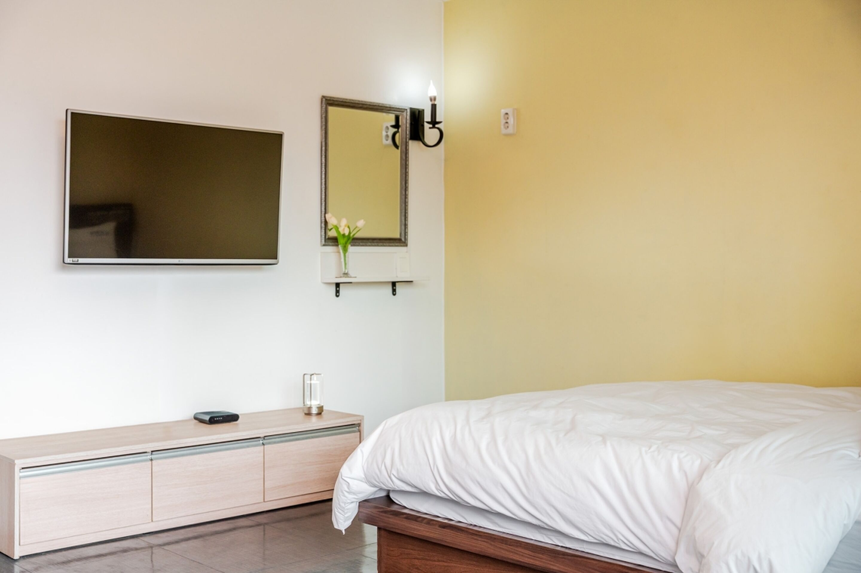 A modern bedroom with a bed, a bedside table, a mirror, and a wall-mounted TV.