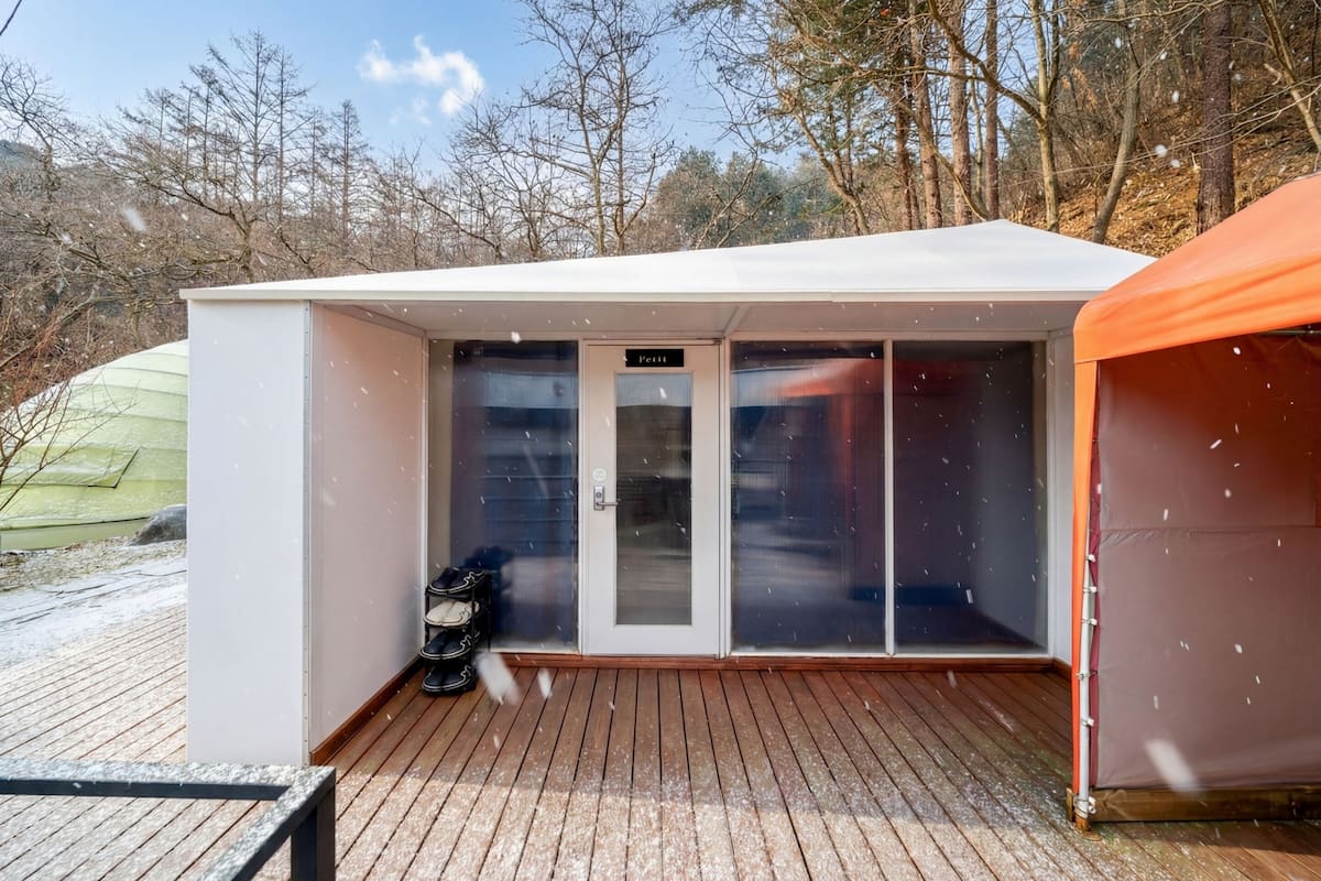 Une petite cabane blanche, composée d’une seule pièce, dotée d’une porte coulissante en verre, d’une terrasse en bois et d’une structure ressemblant à une tente, située sur la droite.