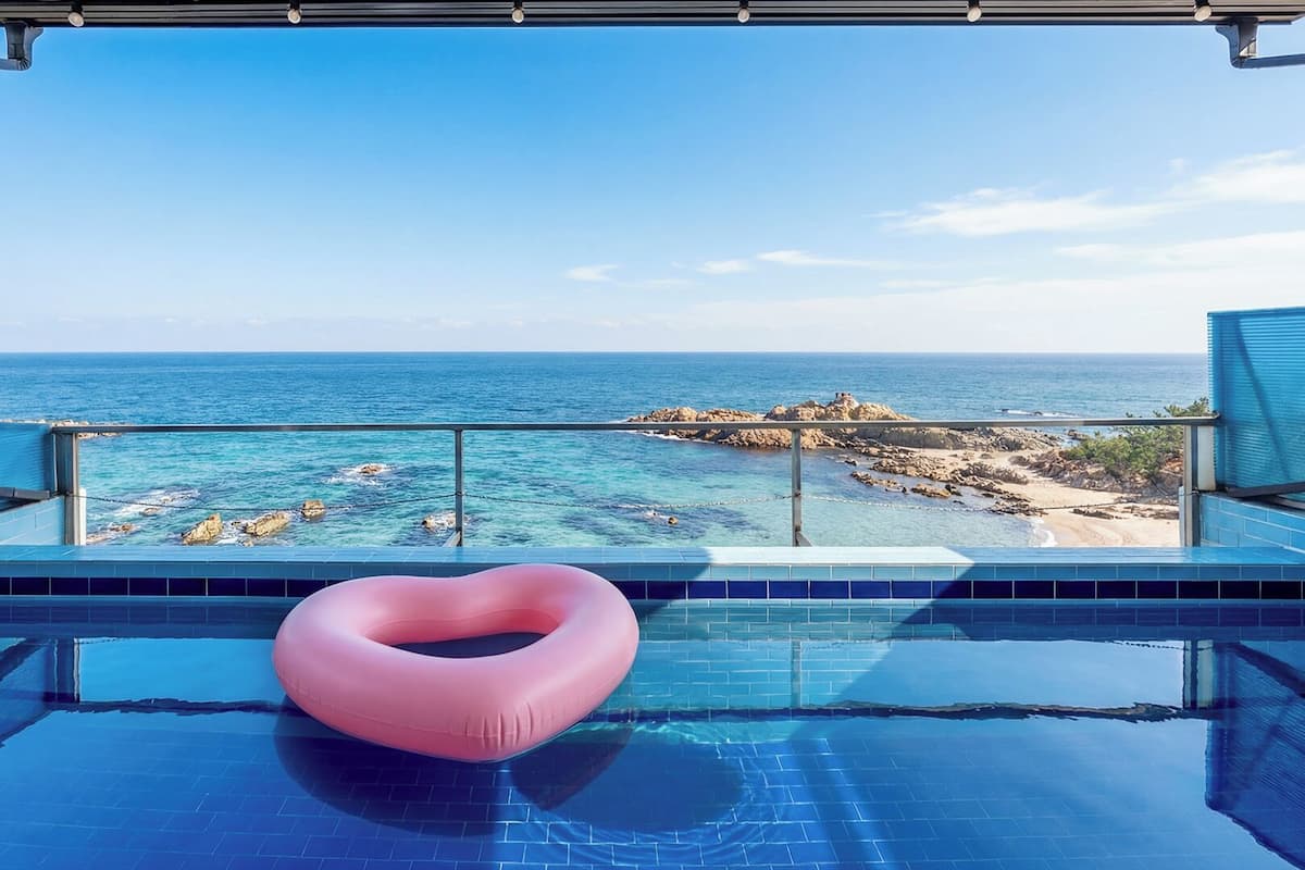 Une piscine avec un flotteur en forme de cœur, une eau d’un bleu cristallin et une vue sur l’océan et la côte rocheuse.