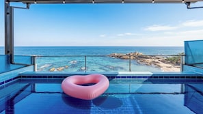En pool med en hjärtformad badring, kristallklart blått vatten och utsikt över havet och den steniga kustlinjen.