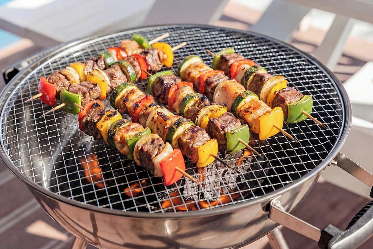 Un barbecue extérieur rond, en acier inoxydable, avec des brochettes de viande et de légumes.