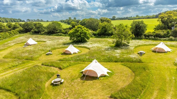 A campsite with multiple tents arranged in a circular pattern on a grassy field, surrounded by trees and open fields. - Glamping accommodation in Norfolk, Blancas Bell Tents sleeps up to 20 people. (Ringstead)