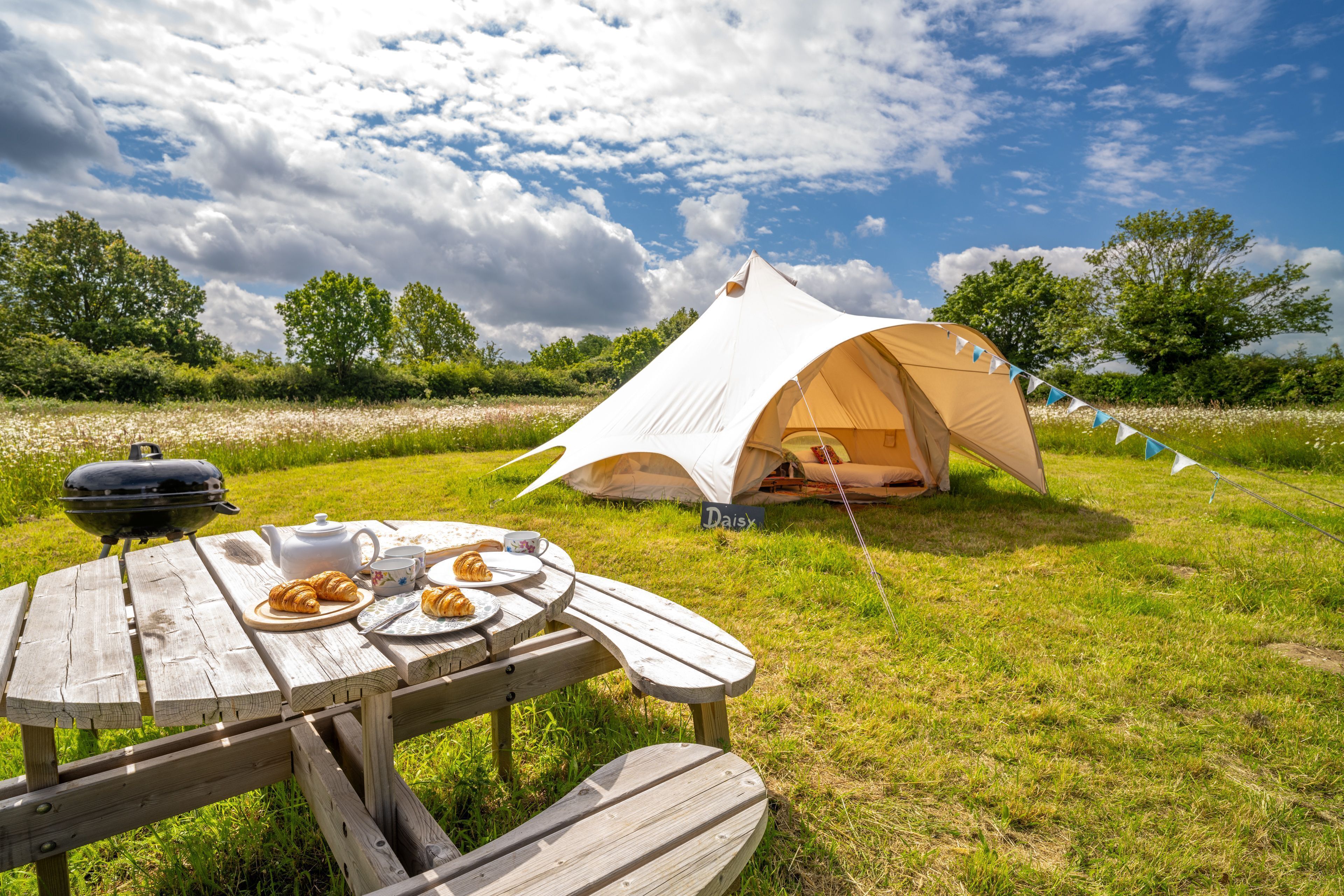 A picnic setup with a tent labeled "Daisy," a wooden table with croissants, a kettle, and a plate of food.