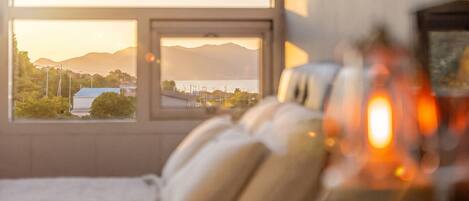 A room with a bed, pillows, a window, and a view of mountains and a body of water.