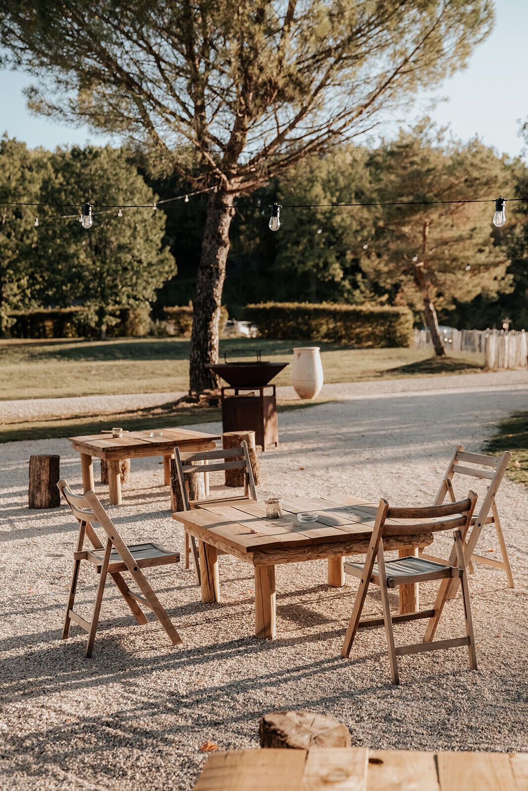 An outdoor seating area with wooden tables and chairs, a large tree, and a gravel ground.