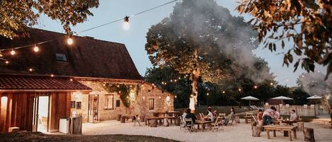 Un espace extérieur aménagé pour les repas, avec des bâtiments en pierre, des guirlandes lumineuses et un grand arbre.