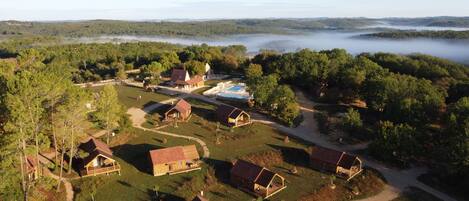 An aerial view of a resort with multiple cabins, a swimming pool, and a forested backdrop.
