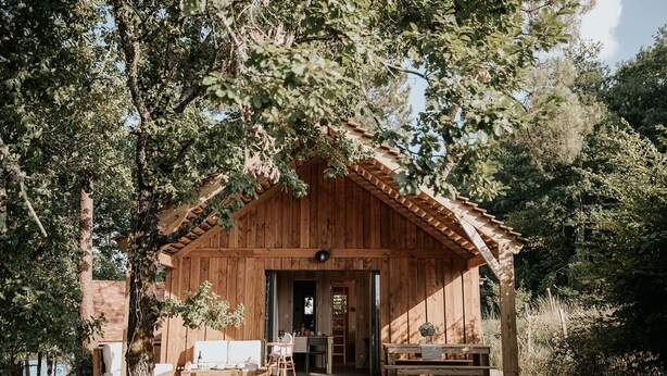 A rustic wooden cabin with a porch, a dining area, and a kitchen visible through the open door. - Domaine Saint-Amand (Saint-Amand-de-Coly)