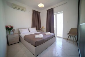 A modern hotel room with a large bed, a bedside table with a lamp, a chair, and a view of the outside through a large window. - Villa Thea (Moires)