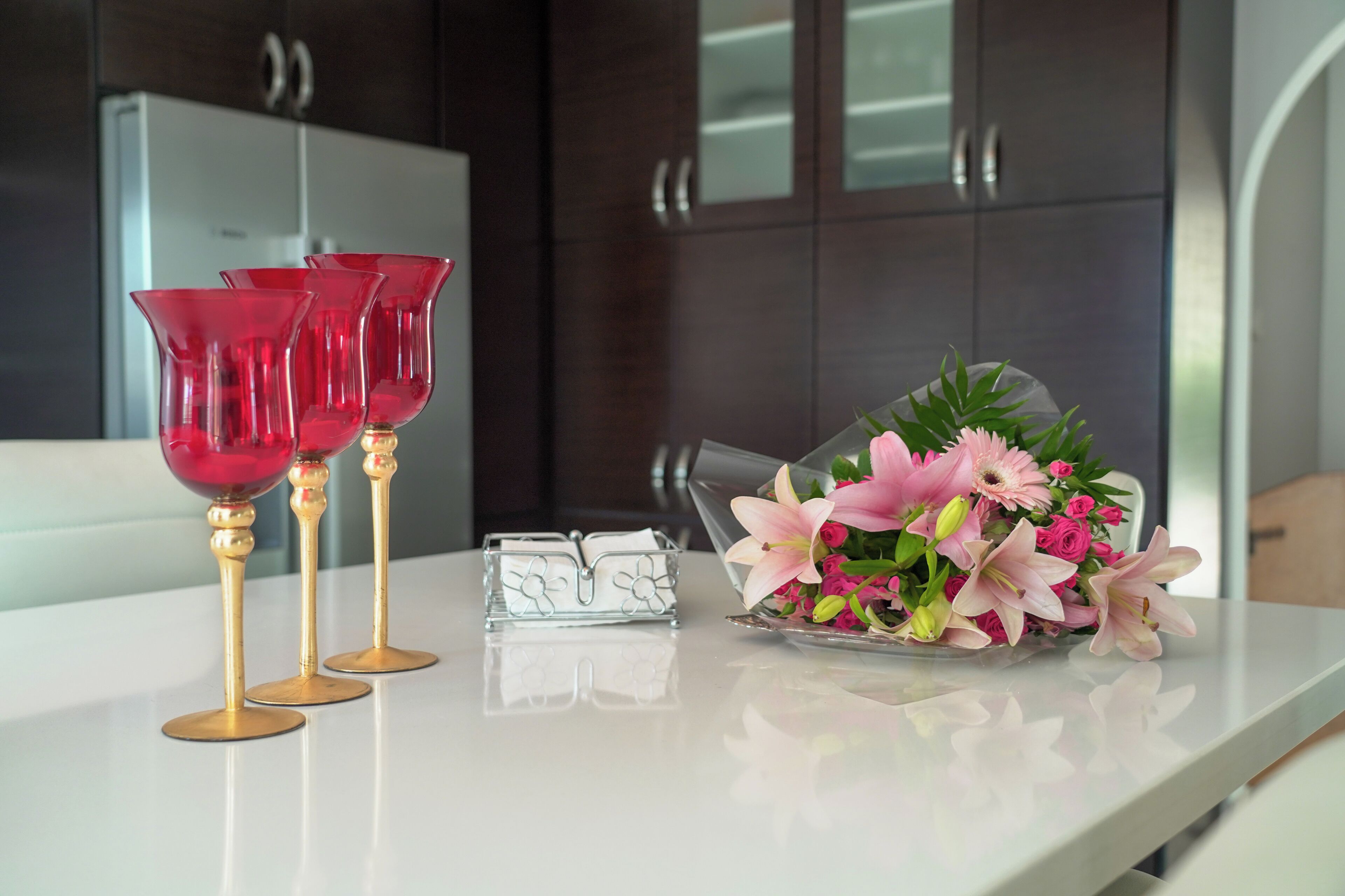 A white table with three red glass cups, a clear glass vase with pink flowers, and a clear glass vase with green leaves.