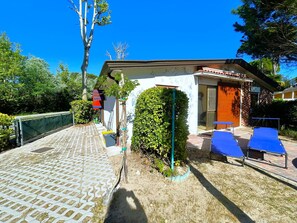 A building with a white facade, a covered outdoor area with blue chairs, and a paved walkway. - VILLA DUNA NORTH 247 - TYPE B1 4+0 - B02477 (Bibione Pineda)