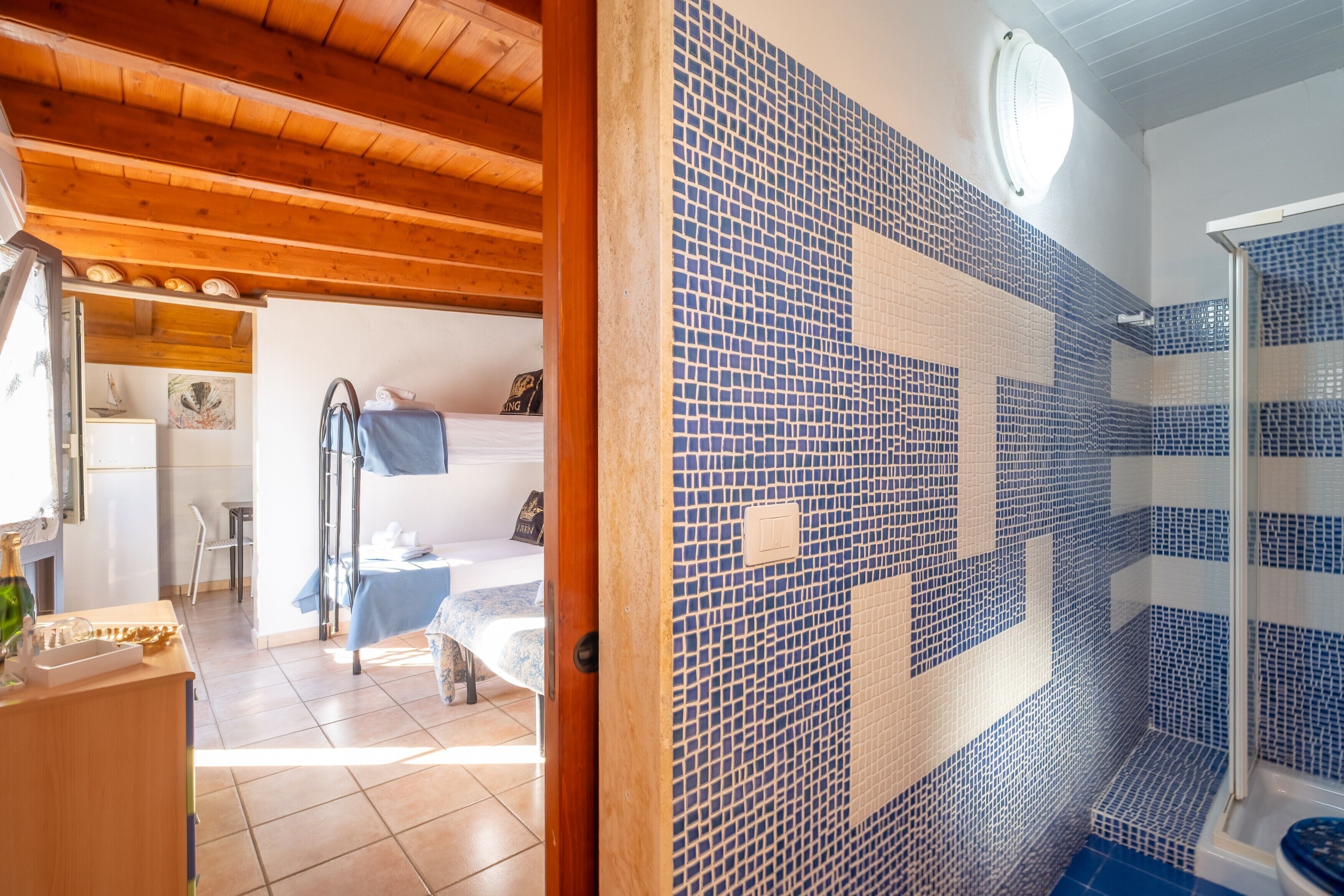 Ein Zimmer mit Holzboden, einem Bett mit blauer Bettdecke, einem kleinen Tisch mit einer Flasche und einem Badezimmer mit blauen Mosaikfliesen.