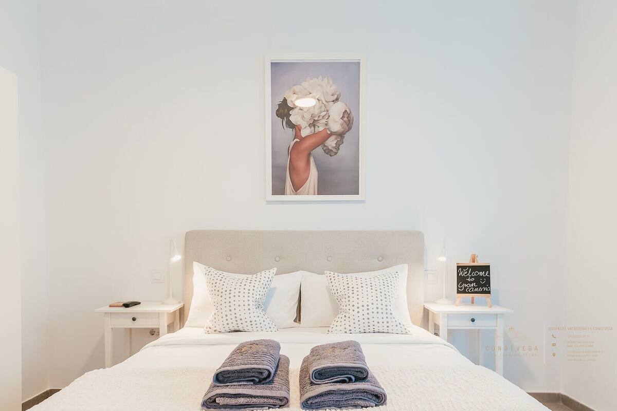 A neatly made bed with two pillows, a framed artwork above, and a small table with a sign.