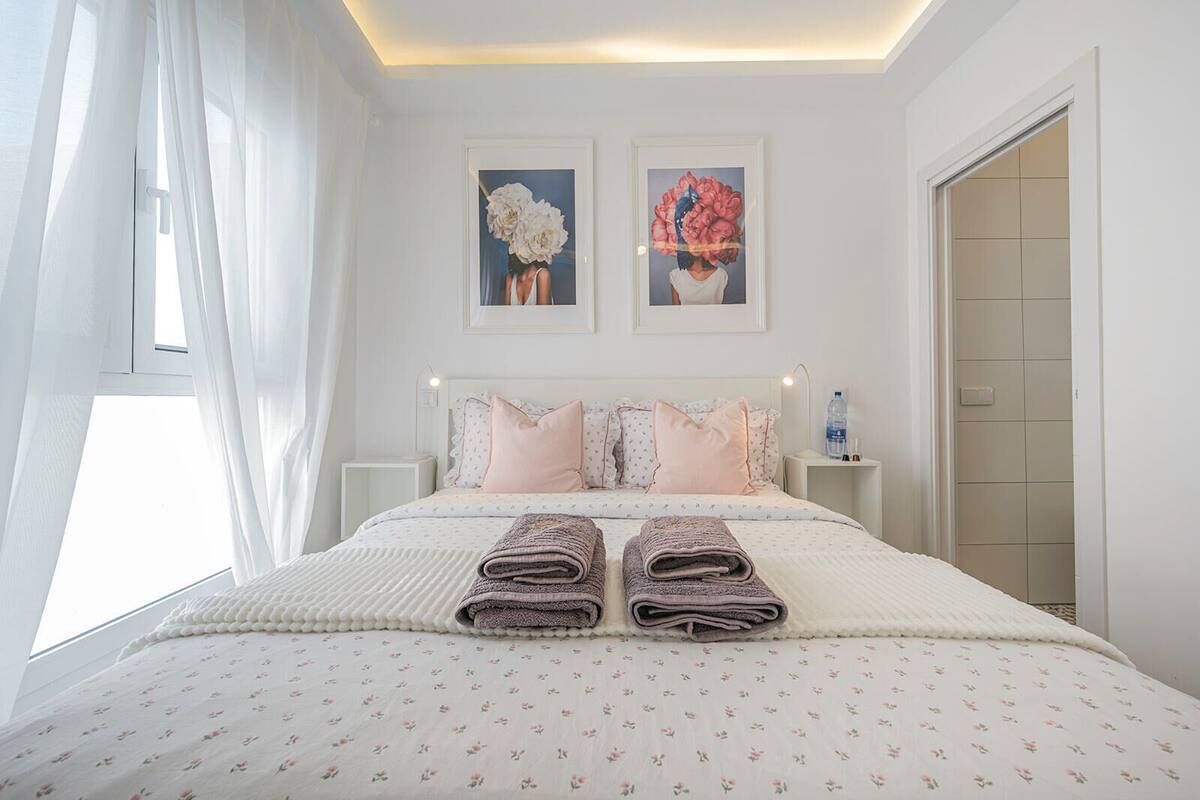 A neatly made bed with a floral-patterned bedspread, two pillows, and folded towels. There are two framed artworks above the bed, a water bottle on a bedside table, and a window with sheer curtains.