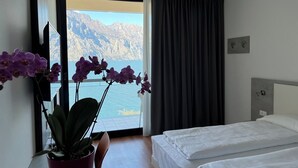 A hotel room with a bed, a potted plant, a window with a view of mountains, and a curtain. - WIND HOTEL (Malcesine)