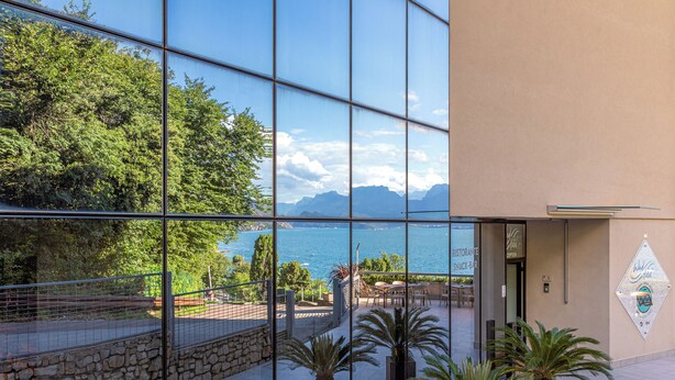 A modern building with large glass windows reflecting a scenic view of a lake and mountains. - WIND HOTEL (Malcesine)