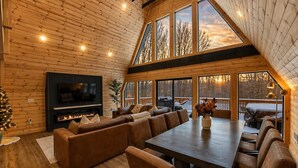 A cabin interior with a large wooden ceiling, a fireplace, a TV, a sofa, and a dining table. - Iconic A-Frame | Hot Tub | Fire Pit | Walk to Lake | Near Ski Resorts (Blakeslee)