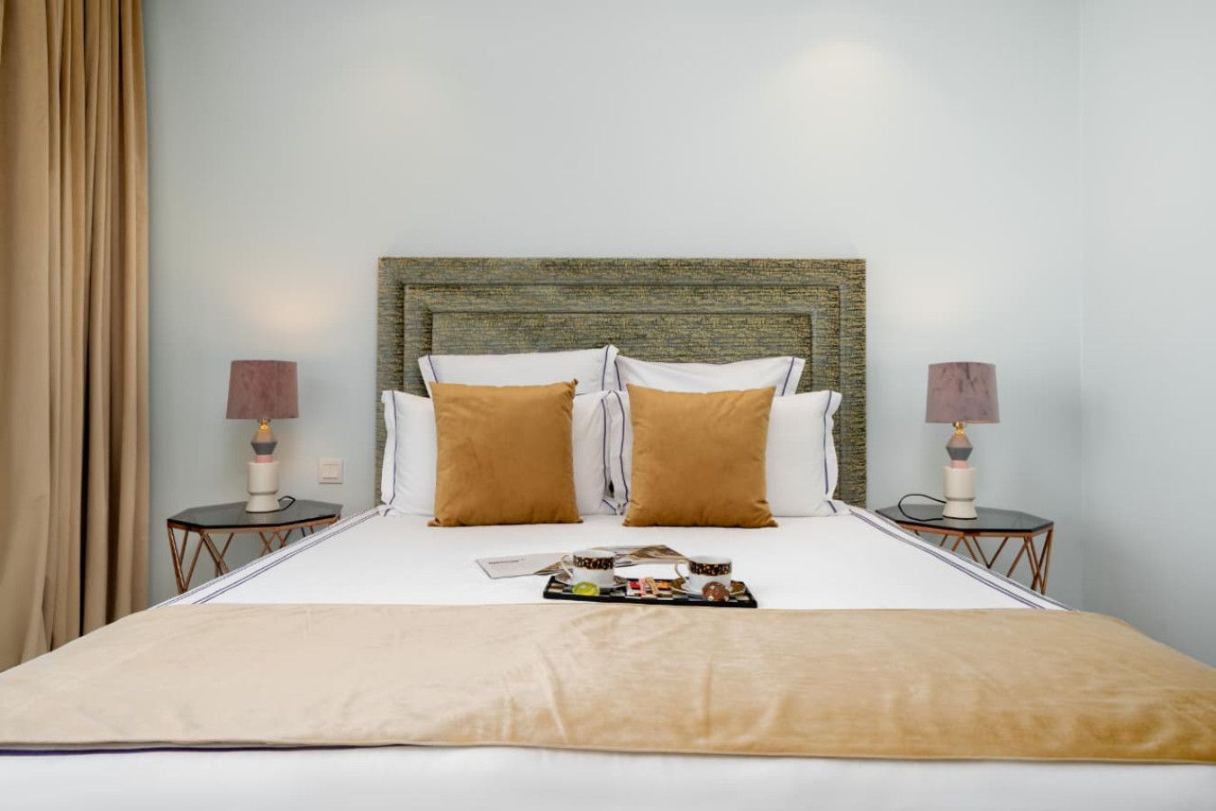 Un lit bien fait, recouvert d’une couvre-lit beige, avec deux oreillers marron et une tête de lit à motifs texturés. On y trouve deux tables de chevet avec des lampes, et un plateau contenant des éléments pour le petit-déjeuner est posé sur le lit.
