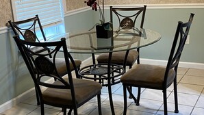 A dining area with a round glass table, four chairs, a wall clock, and a window with blinds. - Welcome to Steve & Tracie’s Gulf Retreat!  (Gautier)