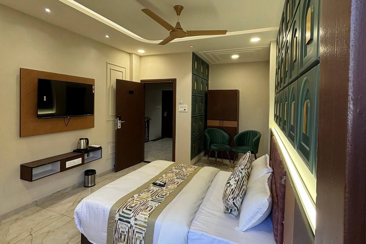 A modern hotel room with a large bed, a ceiling fan, a flat-screen TV, a minibar, and a green chair.