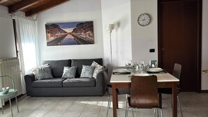 A dining area with a wooden table set for four, a grey sofa with patterned cushions, a large cityscape painting on the wall, and a clock.